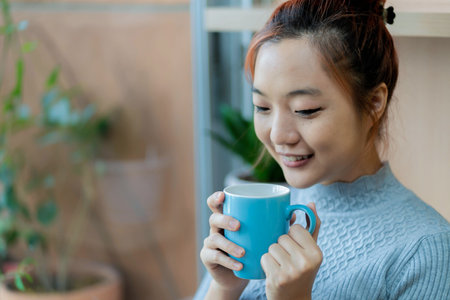 Asian woman smiling softly while holding a blue coffee mug, enjoying a warm beverage and relaxing indoors with natural light and a peaceful atmosphereの写真素材