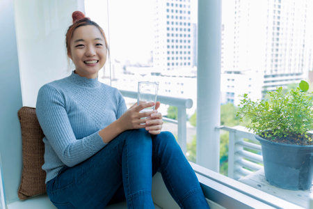 Smiling Asian woman sitting by a large window holding a glass of water, relaxing in a cozy urban apartment with city view, enjoying a moment of wellness and happinessの写真素材