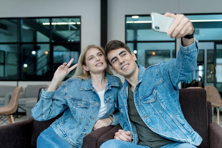 Smiling young couple taking a selfie together in a modern lounge, wearing matching denim jackets and making peace sign for a cheerful social media photoの写真素材