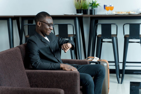 Businessman in suit checking time on wristwatch while waiting with suitcase in modern lounge, travel preparation and punctuality in professional settingの写真素材