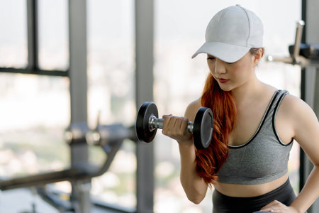 Focused woman lifting dumbbell during strength training workout at modern gym, wearing fitness...