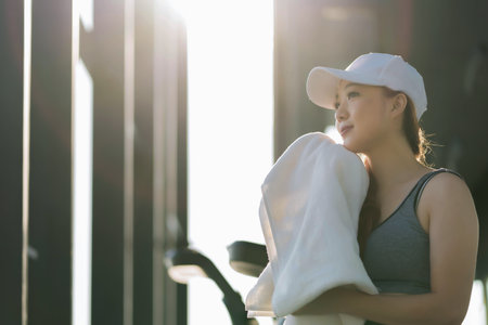 Fit woman in gym clothes resting with towel on shoulder, looking peaceful in warm morning sunlight...
