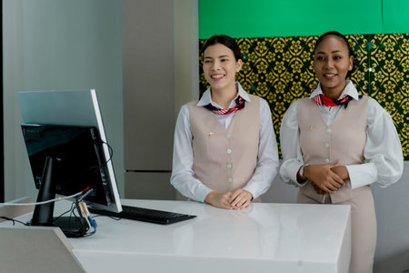 Two friendly female airline staff in uniform standing at airport check-in counter, ready to assist passengers with customer service and flight check-in processの写真素材