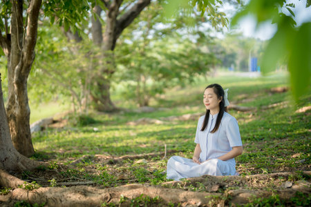 Asian woman meditating under green trees in peaceful park environment, practicing mindfulness and...