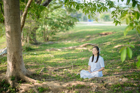 Young woman practicing meditation in nature under tree shade, peaceful atmosphere in green park, mindfulness and relaxation for spiritual and mental well-beingの写真素材