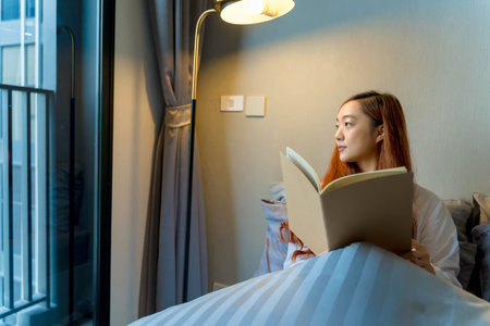 Young woman reading a book while sitting comfortably in bed near window with soft lighting, creating a cozy and peaceful morning atmosphere in a modern roomの写真素材
