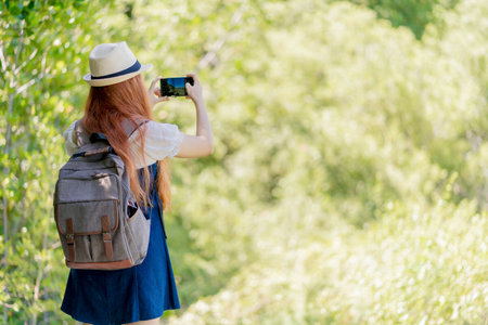 Rear view of young woman with backpack and hat photographing nature with smartphone during hike on a sunny day in lush green forest trail.の写真素材