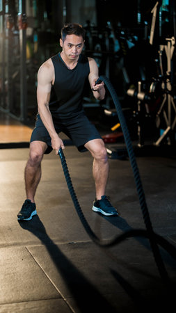 Focused young man performing battle rope workout in a dark gym, showcasing strength, endurance, and intense functional training for full-body conditioningの写真素材