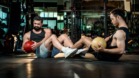 Two men training abs with medicine balls in a gym, seated on the floor and performing synchronized Russian twists for core strength and endurance.の写真素材