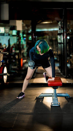 Athletic woman with green hair doing one-arm dumbbell row on bench in gym, focusing on upper body strength and fitness training under dramatic lightingの写真素材