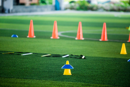 Cone markers is soccer training equipment on green artificial turf with blurry kid players training background. Material for training class of football academyの写真素材