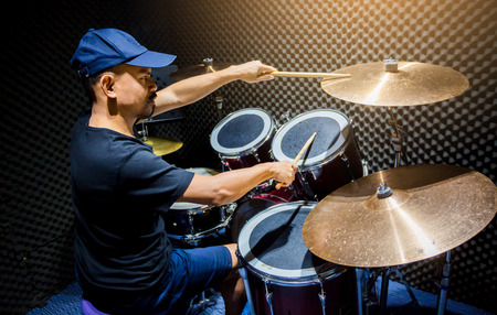 man put black t-shirt and blue cap to playing the drum set with wooden drumsticks in music room , the concept of musical instrumentの写真素材