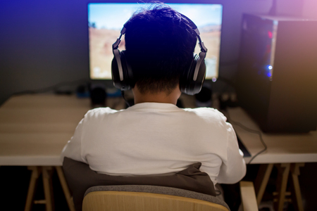 Portrait of asian boy gamer playing games on computer in the room at home, wearing headphones and using backlit colorful keyboard on wooden table.の写真素材