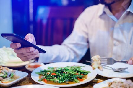 man is eating along with playing smartphone. Lifestyle in today's society. man relaxing having  in the diner and watching some funny videos by smartphone while eating food. man playing games on mobileの写真素材