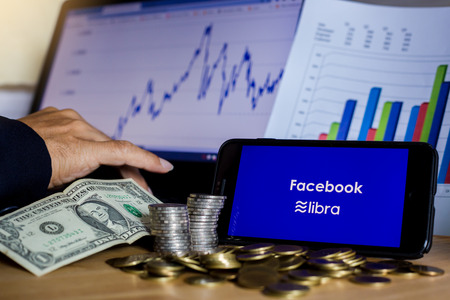 BANGKOK, THAILAND, JULY 23, 2019: Facebook and Libra text display on smartphone with gold silver coin, Dollar bill and hand on laptop background. The concept of business information on technology.のeditorial素材