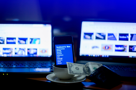 BANGKOK, THAILAND, JULY 10, 2019 : Smart phone display Facebook Libra text and it is in coffee cup with blurry Dollar bill and laptop show business news, The concept of business news in morning.のeditorial素材