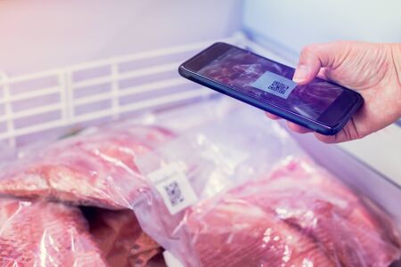 Closeup of woman's hands scanning QR Code of meat products in the freezer with smart phone in...