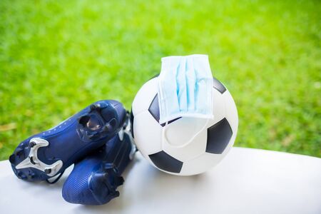 Mask is on football with soccer sport shoes on white floor with green blur background. The concept of Covid-19 impact to football matches.