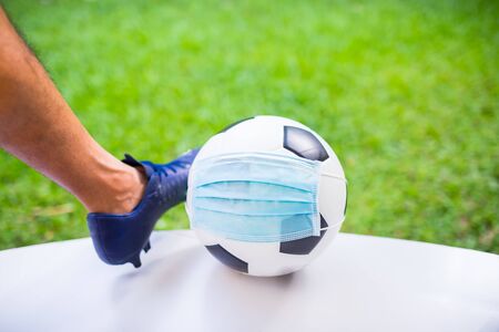 Mask wearing to football with foot and sport shoe of soccer player. The concept of Covid-19 impact ...