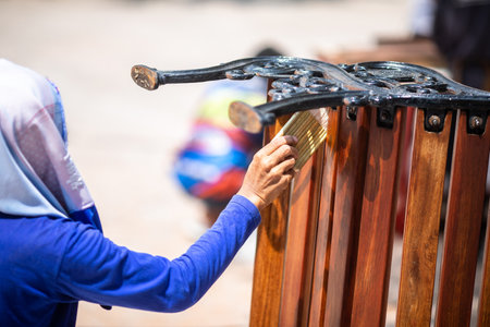 Selective focus to wood with blurry hand of carpenter applying lacquer with a soft paint brush. Hand applies a layer of varnish with a brush on a fragment of a handmade wooden bench.の写真素材