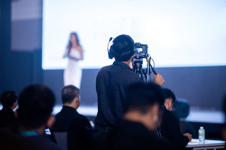 Selective focus to video camera set with blurry cameraman is are  recording to speaker and audience in conference hall or seminar event. exhibition convention center. mass media concept.の写真素材
