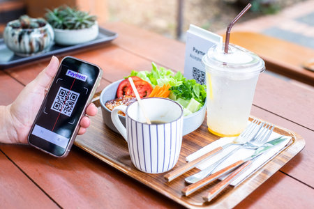 Customer hand using smart phone to scan Qr code payment tag with blur coffee and salad on wooden table to accepted generate digital pay without money. Qr code payment concept.の写真素材