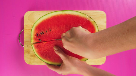top view, female hand cuts fruit with a knife on a wooden board, red watermelon. The concept of...