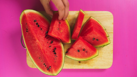 top view, female hand cuts fruit with a knife on a wooden board, red watermelon. The concept of...