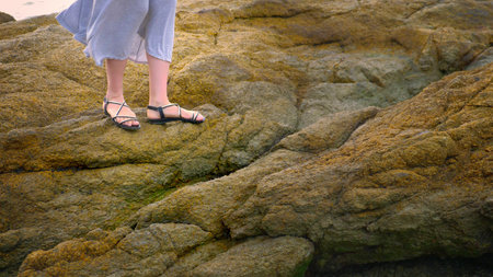 close-up. women's legs in sandals and a long gray skirt are on the rocky seashore at low tideの写真素材