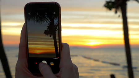 A woman with a mobile phone makes a photo of a tropical beach at sunset.の写真素材