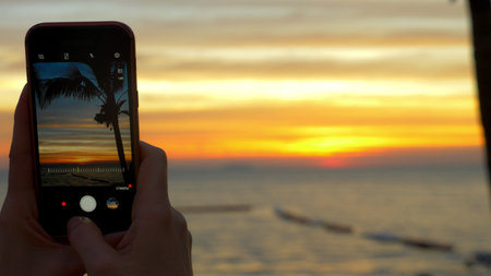 A woman with a mobile phone makes a photo of a tropical beach at sunset.の写真素材