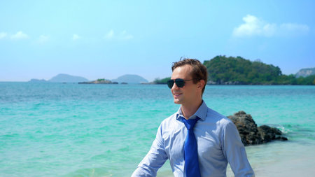 handsome businessman in sunglasses walked along a tropical beach, taking off his tieの写真素材