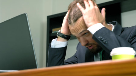 a man in a suit works at a desk in the office on a laptop and drinks coffee from a disposable cup....