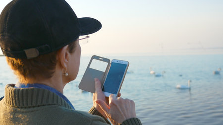 An elderly woman walking on the beach makes a photo of white swans swimming in the sea on her smartphone.の写真素材