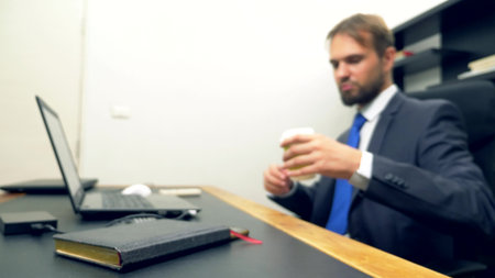 a man in a suit works at a desk in the office on a laptop and drinks coffee from a disposable cup....