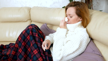 Beautiful woman with a runny nose uses a nasal spray at home, sitting on the sofa in the living room, in a sweater and under a blanketの写真素材
