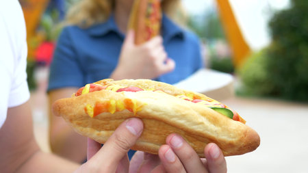 couple young man and woman eating an appetizing hot dog, sitting in the park against the background of walking people, blurred background.の写真素材