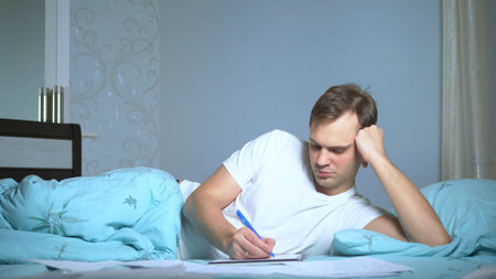 A man writes on paper documents lying in bed. work at home.の写真素材