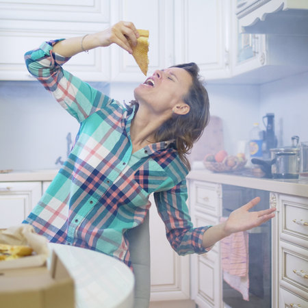 cheerful funny woman eating pizza out of the box at home in the kitchenの写真素材