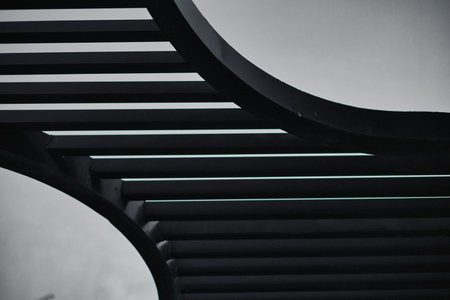 A professional low-angle shot of a modern architectural structure featuring parallel black metal bars and flowing curved steel elements. The geometric pattern creates a striking minimalist silhouette against the clear, deep blue sky of twilight.の写真素材