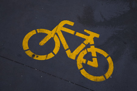 A professional top-down shot of a bright yellow bicycle icon stenciled onto dark, damp pavement. The...