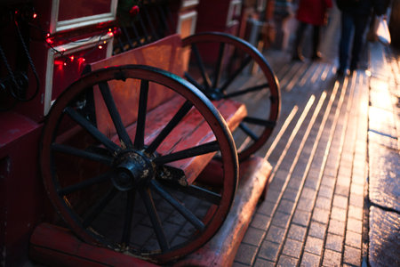 A close-up photograph of a unique outdoor bench designed with oversized black wooden wheels as armrests. The bench and adjacent wall are painted deep red, decorated with small glowing holiday lights. Low-angle sunlight creates long shadows and warm highlights on the wet cobblestone pavement.