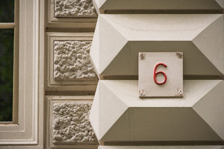 Close up of a vintage house number 6 plaque mounted on an ornate architectural facade. Decorative stone textures, geometric building details and classic address signage create an elegant urban heritage and city architecture concept.