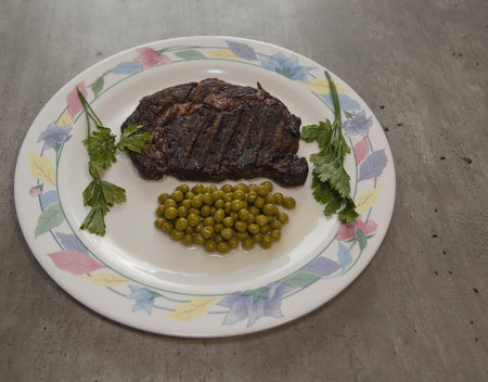 Grilled Rib Eye steak in olive oil and Provencal herbs with green peas and bunches of parsley on a plate with patternsの写真素材