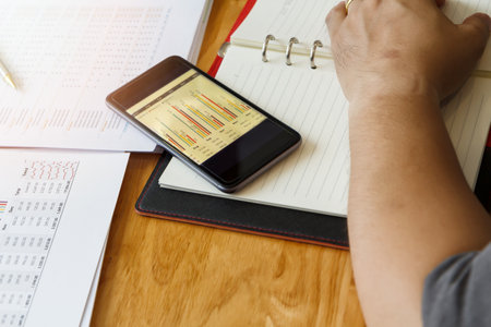 Smartphone with graph near a businessman's arm. Smartphone, diary and report paper on wooden table.の写真素材