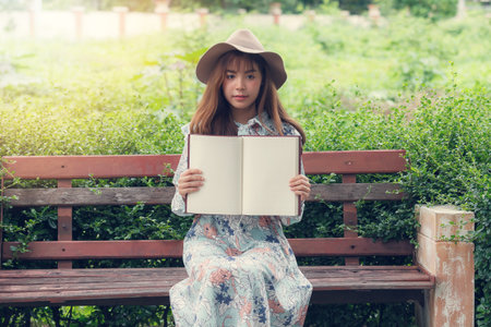 Young asian woman in retro dress sitting on the bench in park. She holding empty blank notebook, showing you something in notebookの写真素材