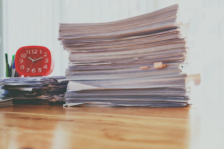 Stack of documents with red clock and office supplies on desk in home office at morning, warm toneの写真素材
