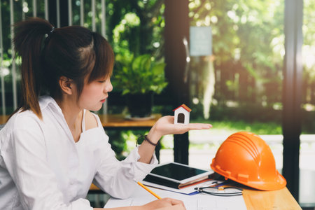 Beautiful young Asian woman holding house model while sitting in front of her working place.の写真素材