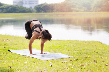 Asian woman practicing yoga doing Crane (Crow) Pose, Bakasana pose on the mat beside a lake in...