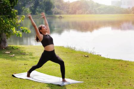 Asian woman practicing yoga doing Warrior I pose, Virabhadrasana I pose on the mat beside a lake in outdoor park.の写真素材